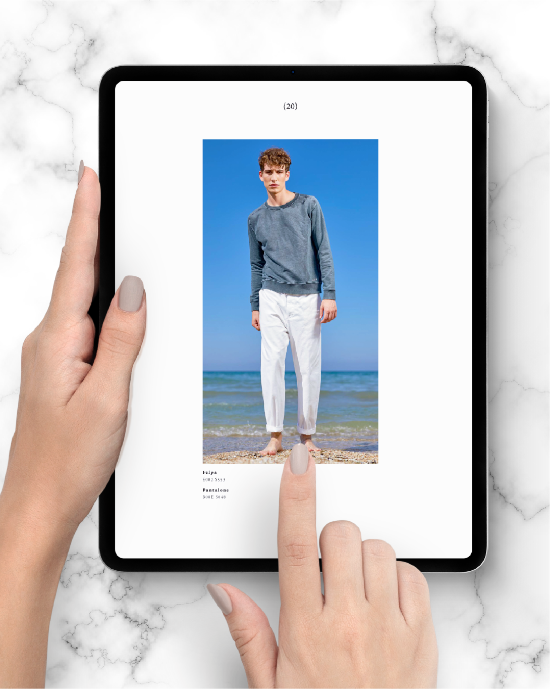 Tablet held by a hand with another hand's finger pointing at the screen showing a full-length image of a young man standing barefoot on a pebbly beach wearing a long-sleeve grey sweatshirt and white pants against a clear blue sky and sea background.