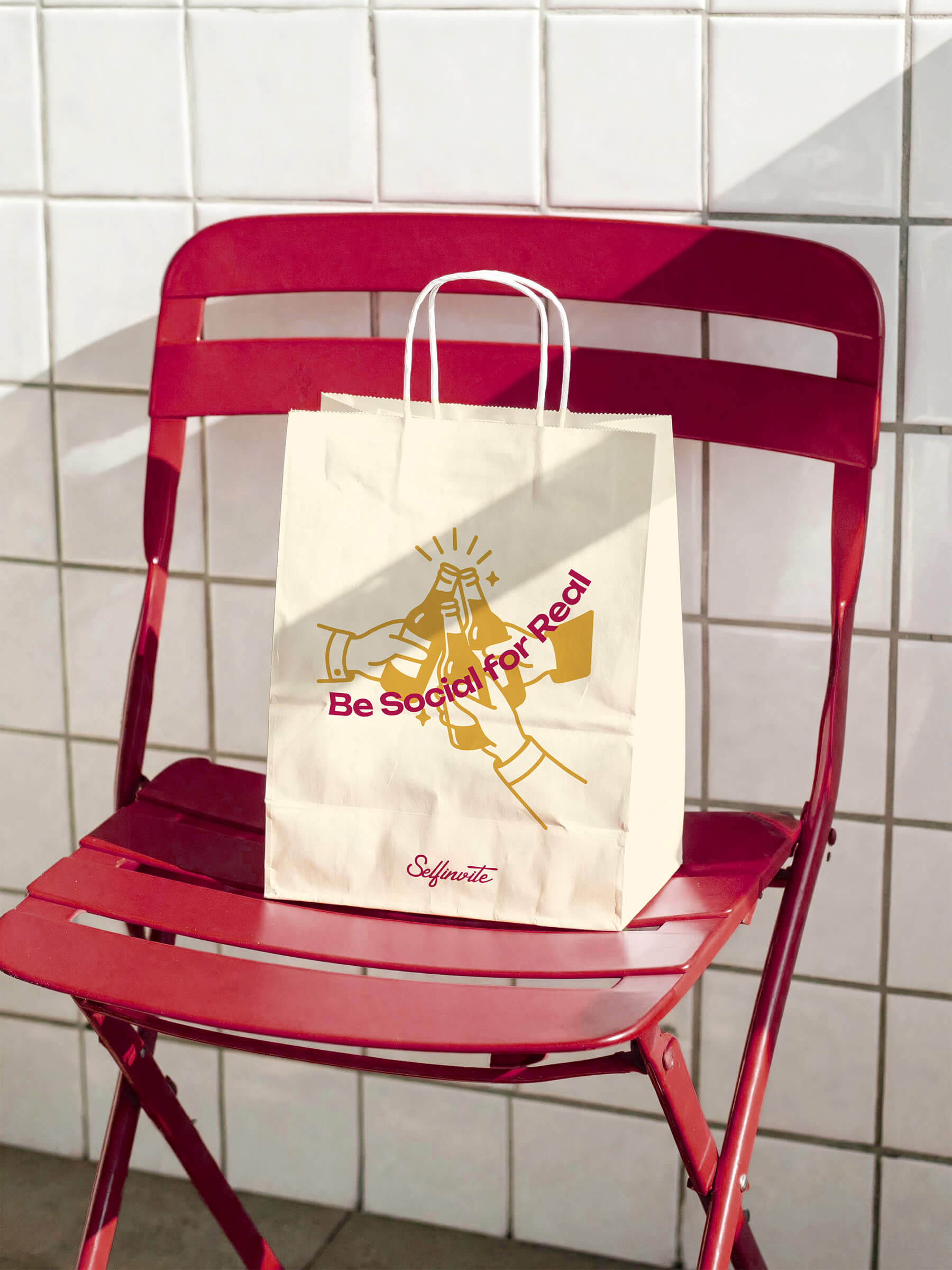 A white paper bag with handles sitting on a red metal folding chair against a white tiled wall, showing an illustration of hands clinking bottles and text that reads 'Be Social for Real' and 'Selfinvite' at the bottom.