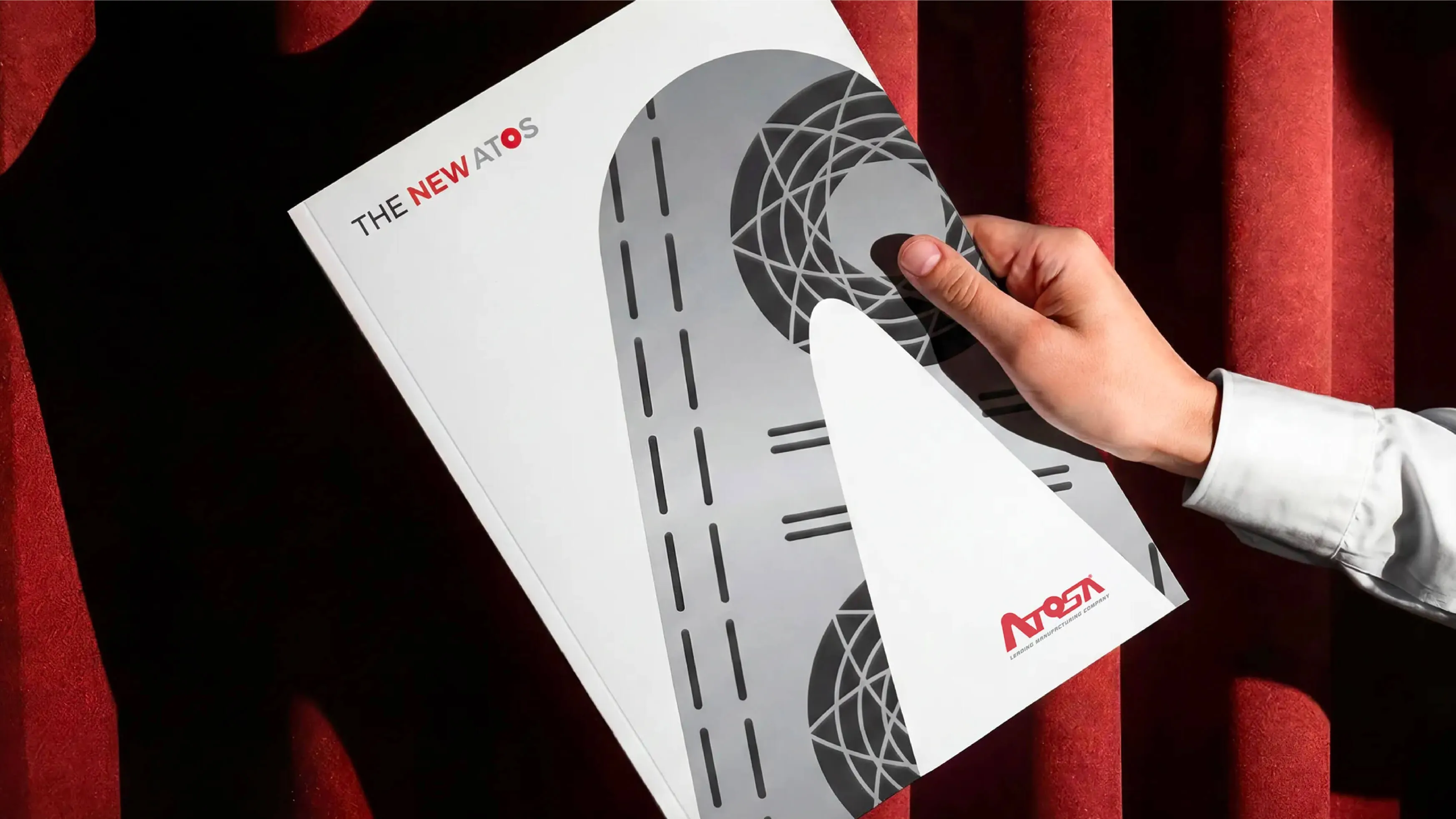 Hand holding a white and gray brochure titled 'THE NEW ATOS' with a geometric design and a red ATOS logo on a red fabric background.