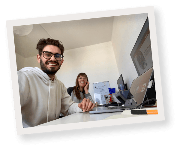 Tommi & Pam smiling at the camera in their office with laptops, monitors, and a whiteboard in the background.