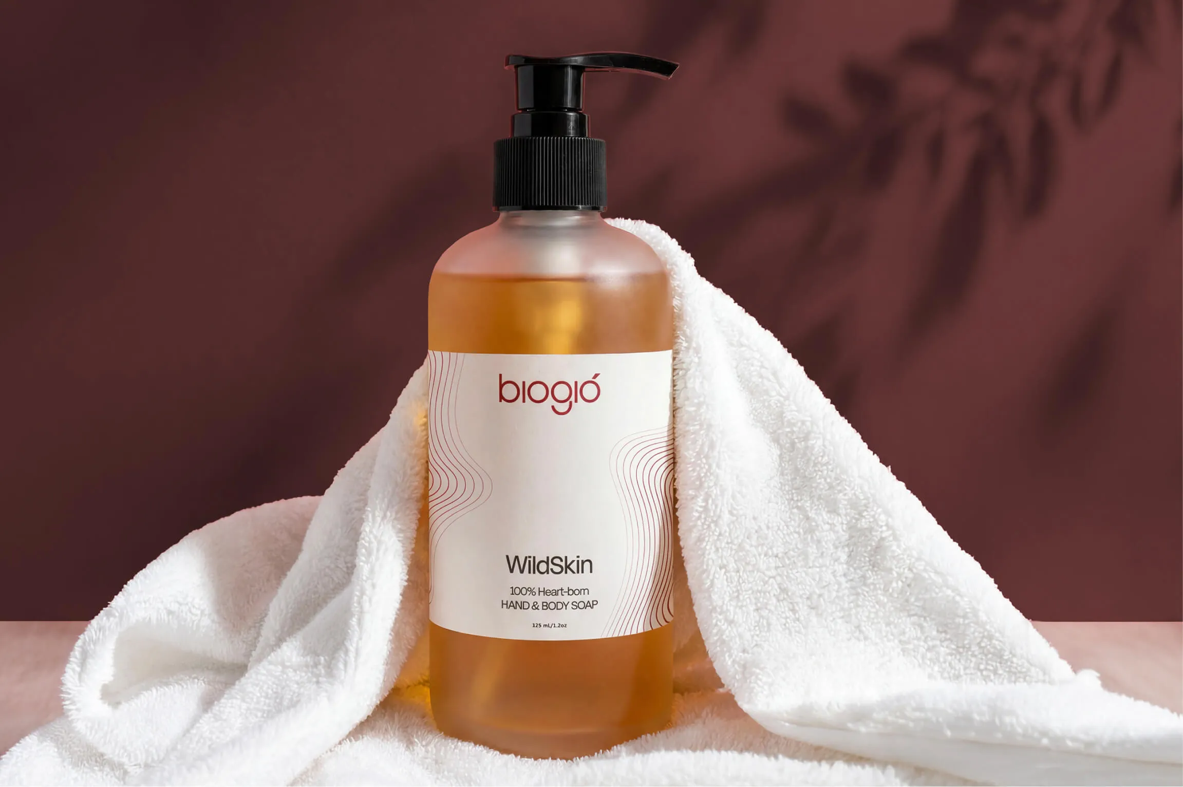 Bottle of Biogio WildSkin hand and body soap with a pump dispenser, amber liquid inside, placed on a white towel with a brown background and shadow of leaves.