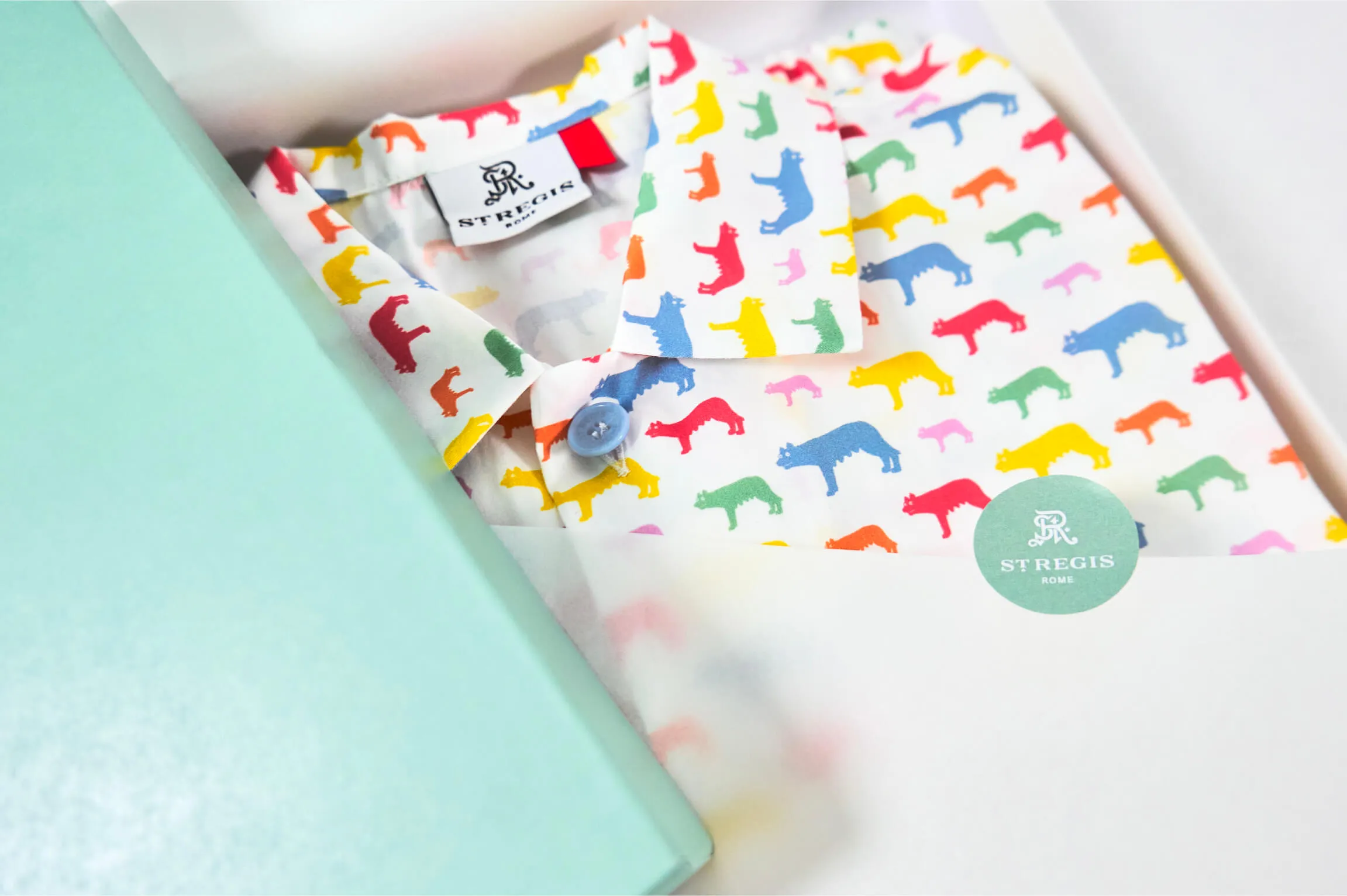 Folded white pajama with a colorful pattern of small cartoon-style wolves in red, yellow, blue, green, orange, and pink, partially wrapped in translucent paper with a green circle sticker labeled 'ST REGIS ROME', next to a light teal box.