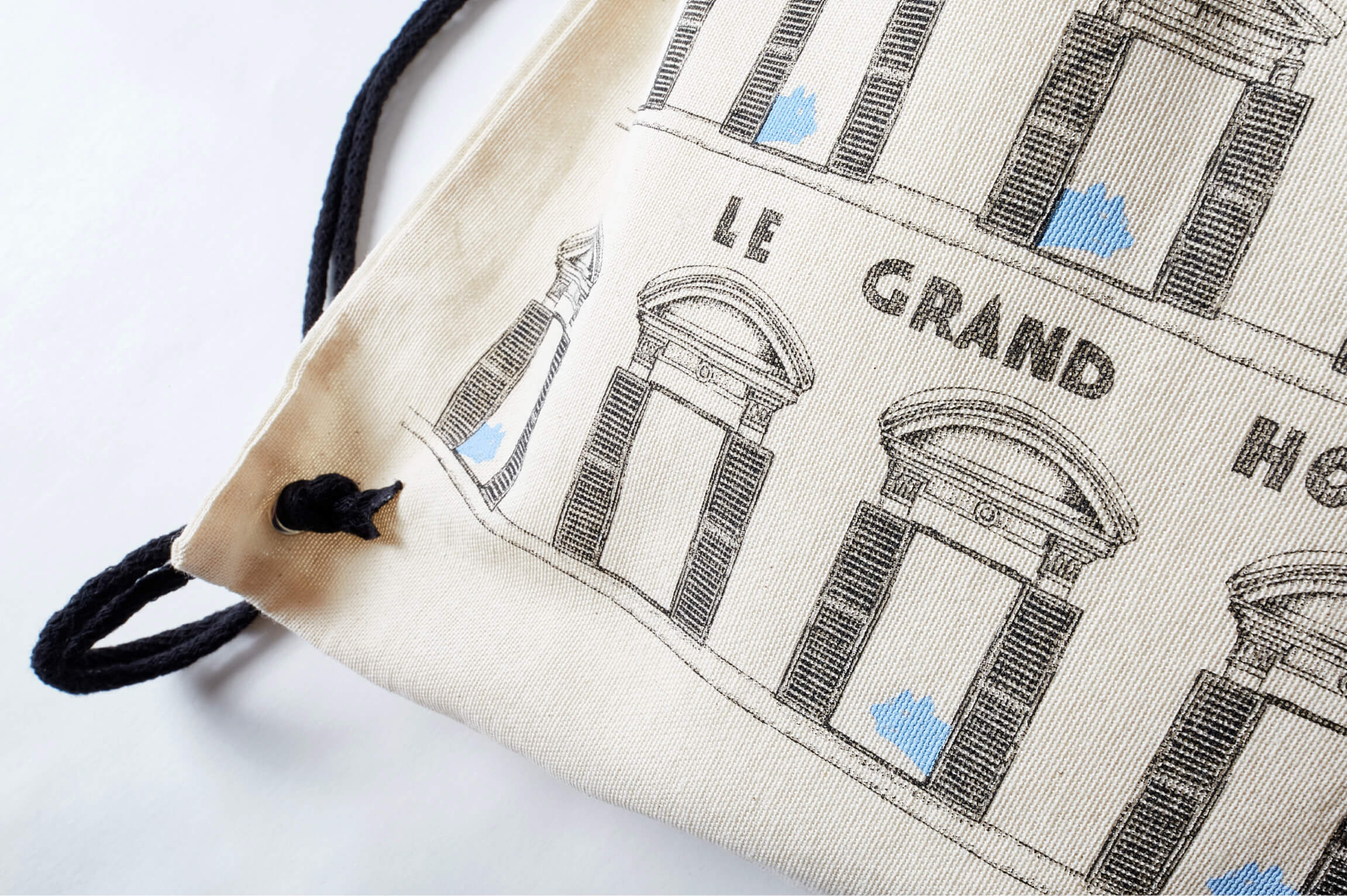 Close-up of a beige canvas drawstring bag with black straps, featuring black architectural sketches and the text 'LE GRAND HOTEL'.