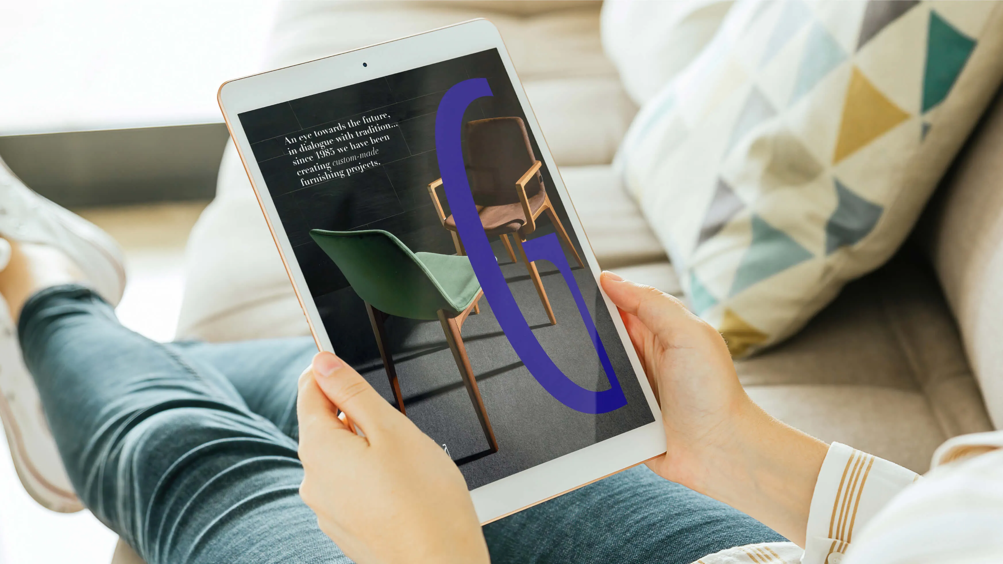 Person holding a tablet displaying an advertisement featuring two modern chairs, one green and one brown, with text about creating custom-made furnishing projects since 1985 and a large stylized purple letter 'G'.