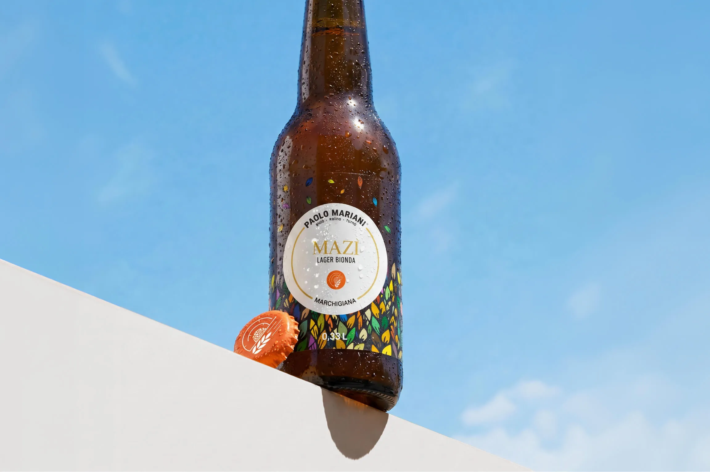 A cold brown bottle of Mazi Lager Bionda beer with condensation droplets, standing on a white surface against a clear blue sky, next to an orange bottle cap.