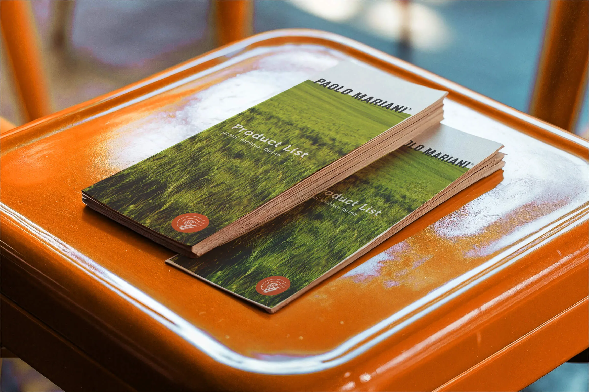 Two stacks of brochures titled 'Product List' with a green grass field cover and the name 'Paolo Mariani' at the top, placed on an orange glossy table.