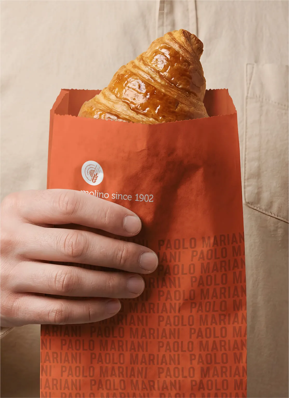 Hand holding a red paper bag with the text 'PAOLO MARIANI' repeated and a croissant peeking out from the top.