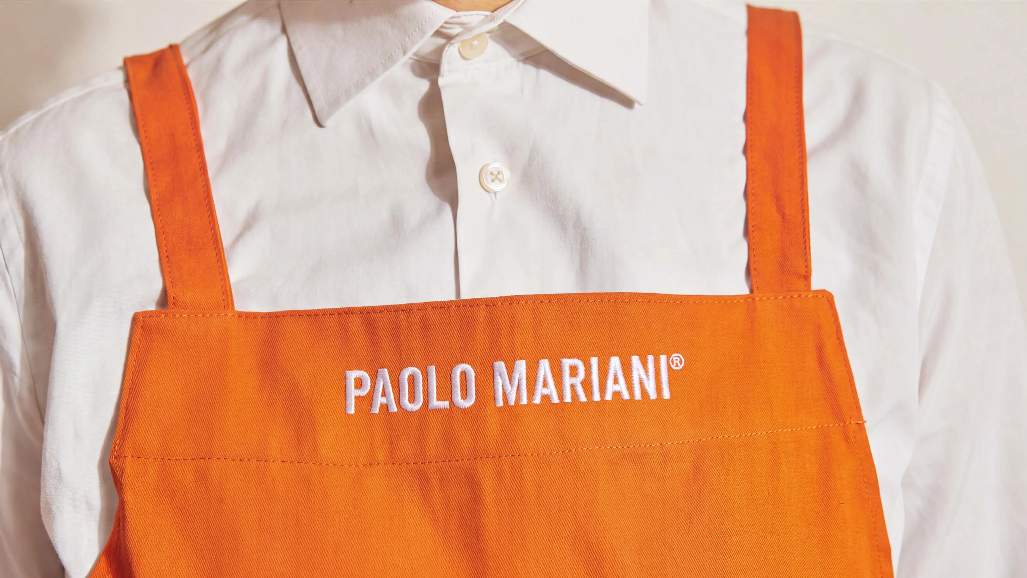 Close-up of a person wearing an orange apron over a white button-up shirt; the apron has 'PAOLO MARIANI®' embroidered in white on the chest.