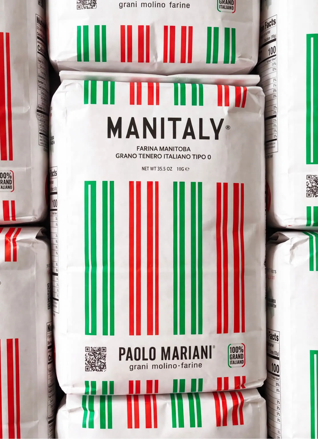 Stacked white bags of Manitaly Farina Manitoba Italian flour with green, white, and red barcode-like stripes, featuring Paolo Mariani branding and 100% Grano Italiano label.