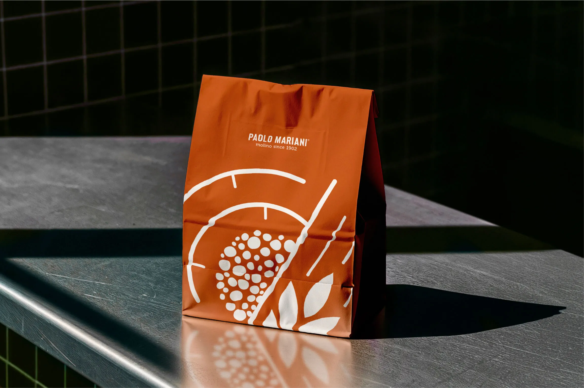 Orange Paolo Mariani branded paper shopping bag with white abstract circular and leaf patterns, standing on a shiny gray metal surface with a shadow cast behind it.