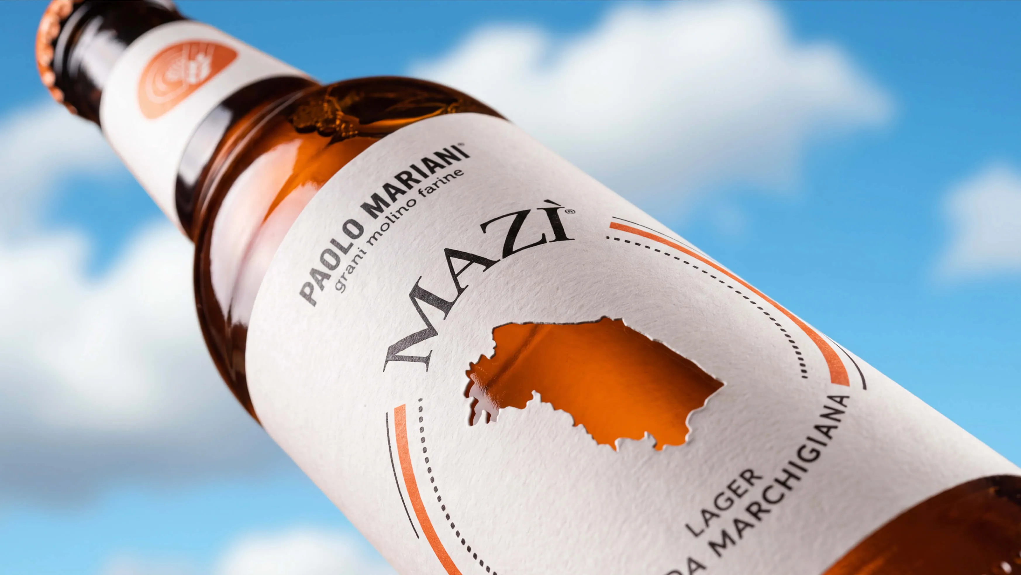 Close-up of a brown beer bottle with a white label featuring the text 'Paolo Mariani grani molino farine' and 'Mazì', with a cut-out shape showing the amber beer inside, and 'Lager Marchigiana' at the bottom against a blue sky with clouds background.