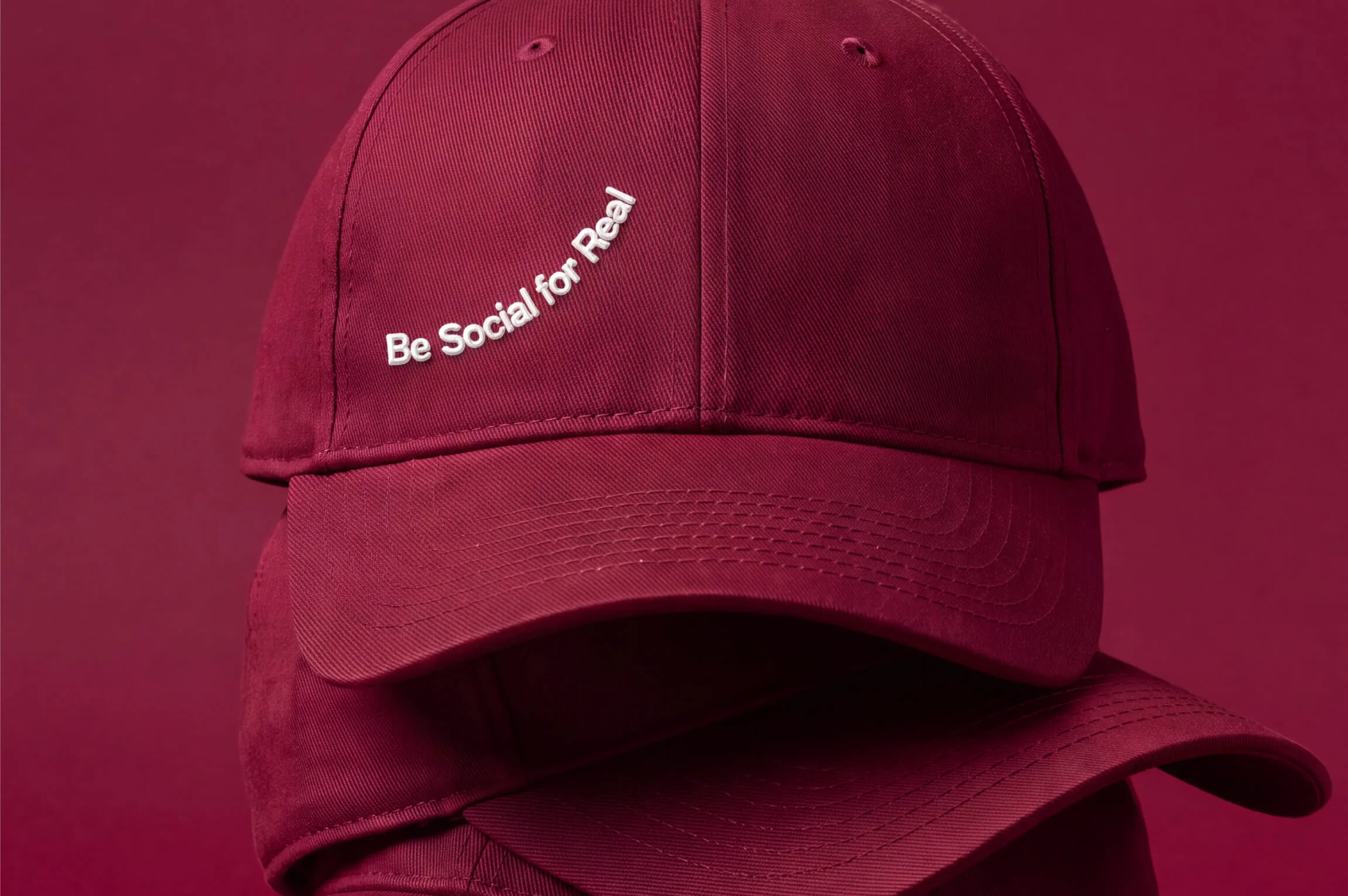 Close-up of two maroon baseball caps stacked, the top one embroidered with white text reading 'Be Social for Real.'