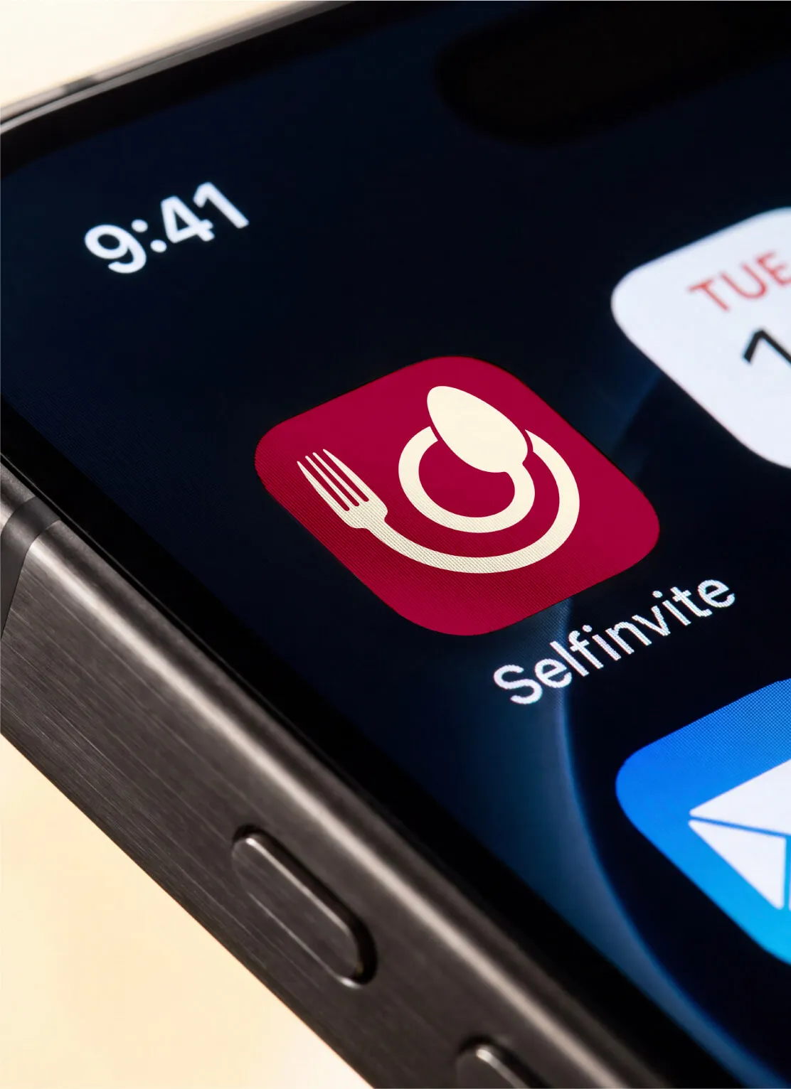 Close-up of a smartphone screen displaying the Selfinvite app icon featuring a fork, spoon, and plate on a red background.
