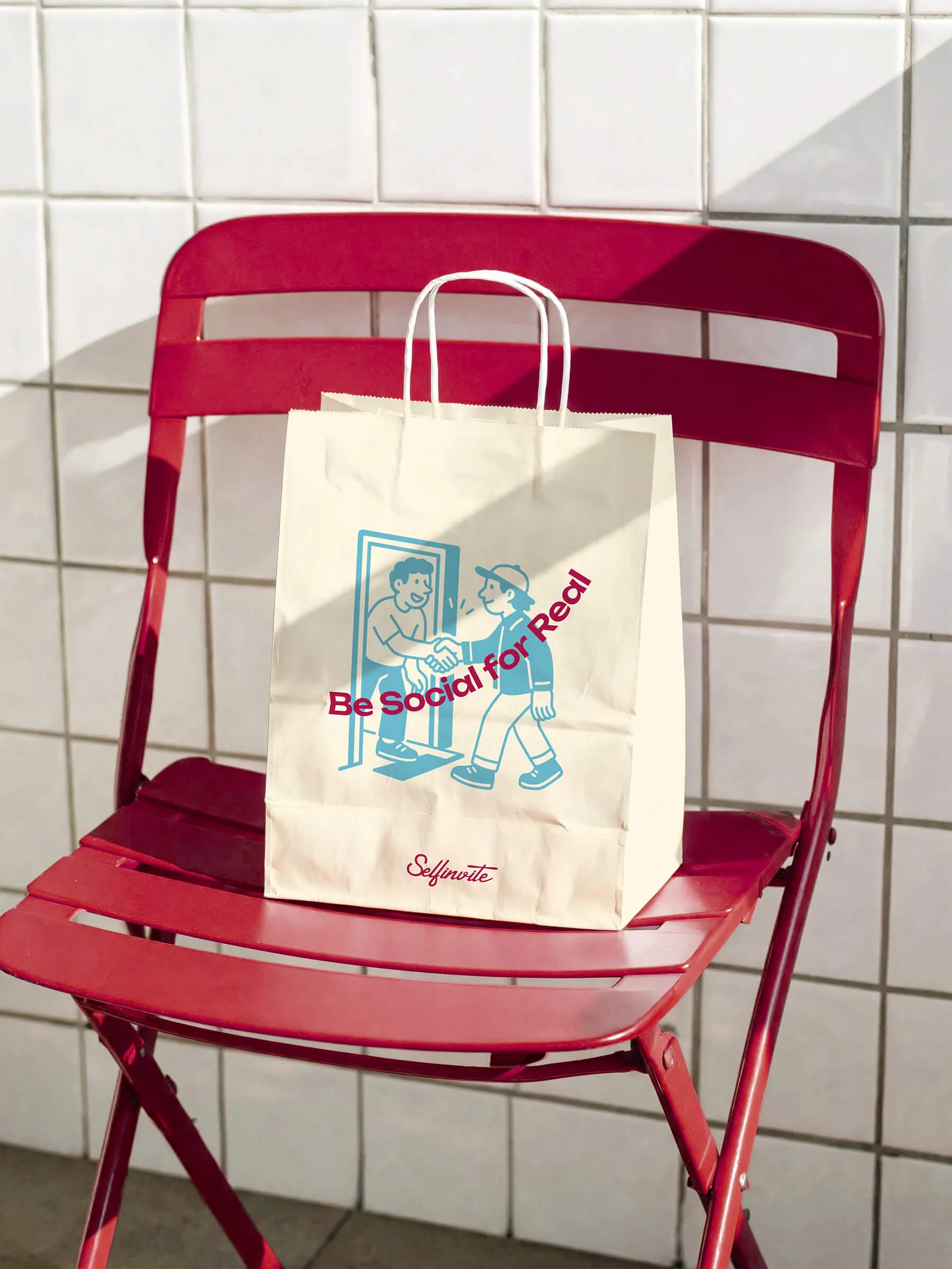 White paper shopping bag with blue illustration of two people shaking hands and the pink text 'Be Social for Real' placed on a red folding chair in front of white tiled wall.
