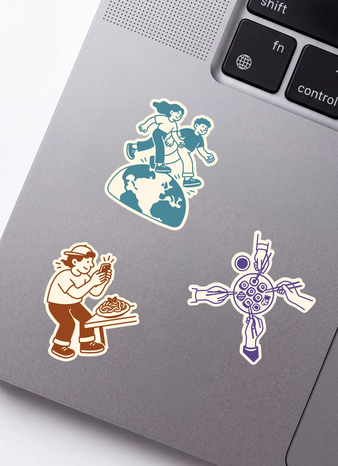 Three illustrated stickers on a laptop showing: a pair of people holding hands and running over a globe, a person taking a photo of a plate of spaghetti, and four hands using chopsticks to eat sushi from a round plate with soy sauce.