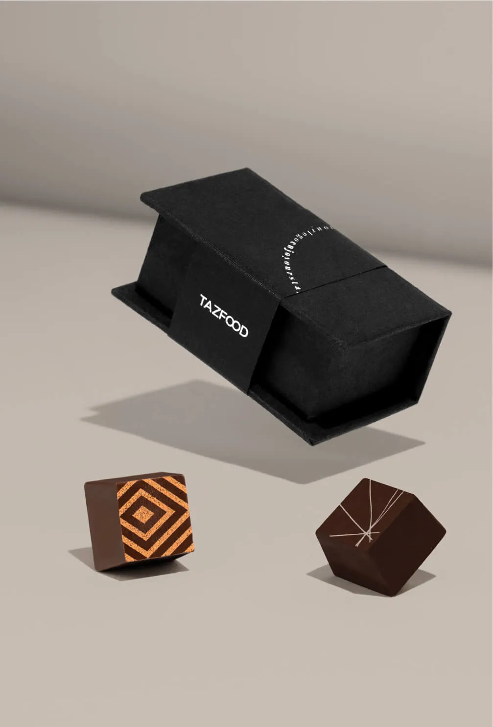 Two chocolates, one with an orange geometric pattern and the other with white line accents, placed near a black rectangular box labeled TAZFOOD on a light beige surface.