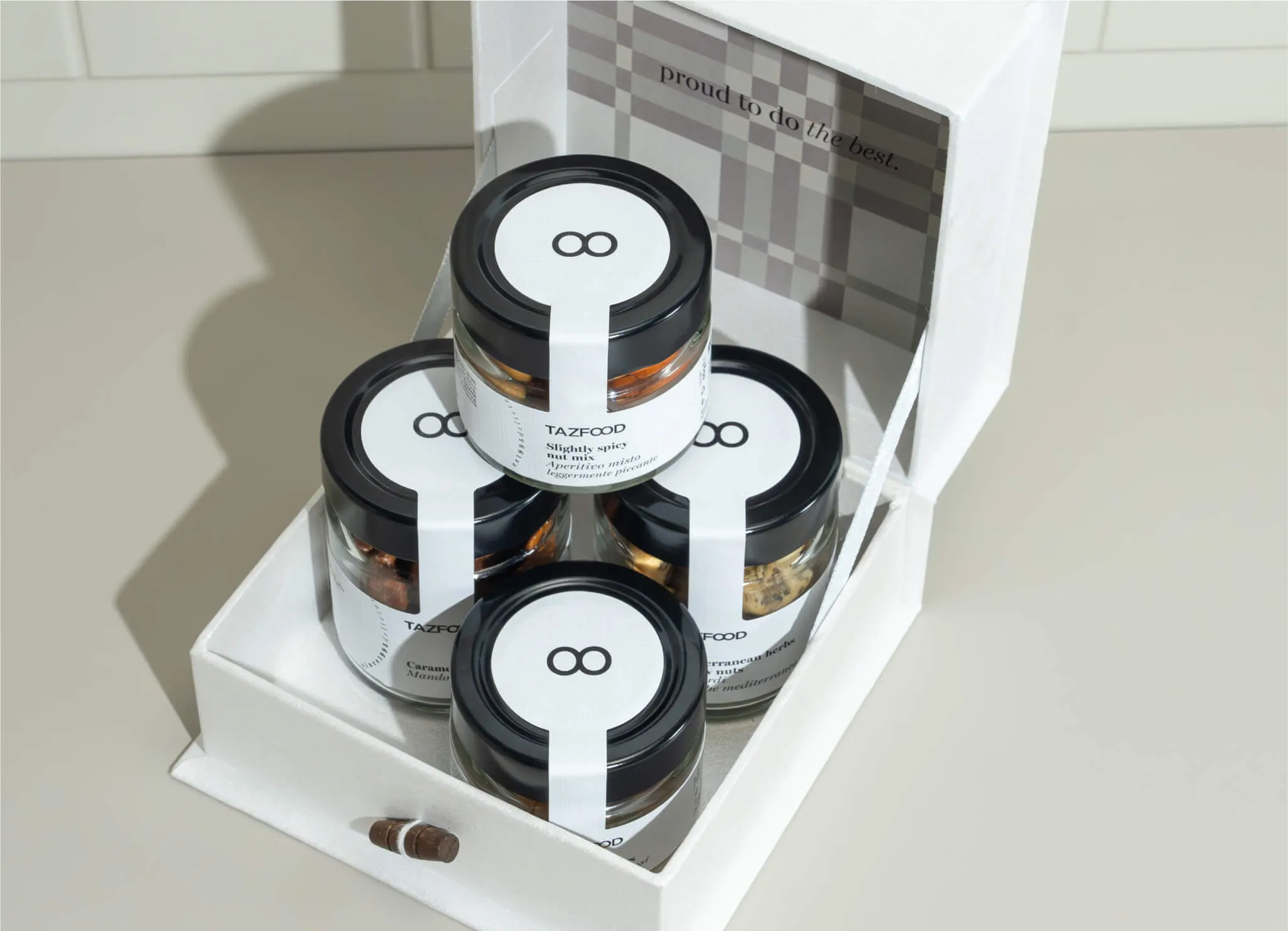 Four glass jars with black lids labeled TAZFOOD inside an open white box with a plaid patterned interior flap that says 'proud to do the best.'