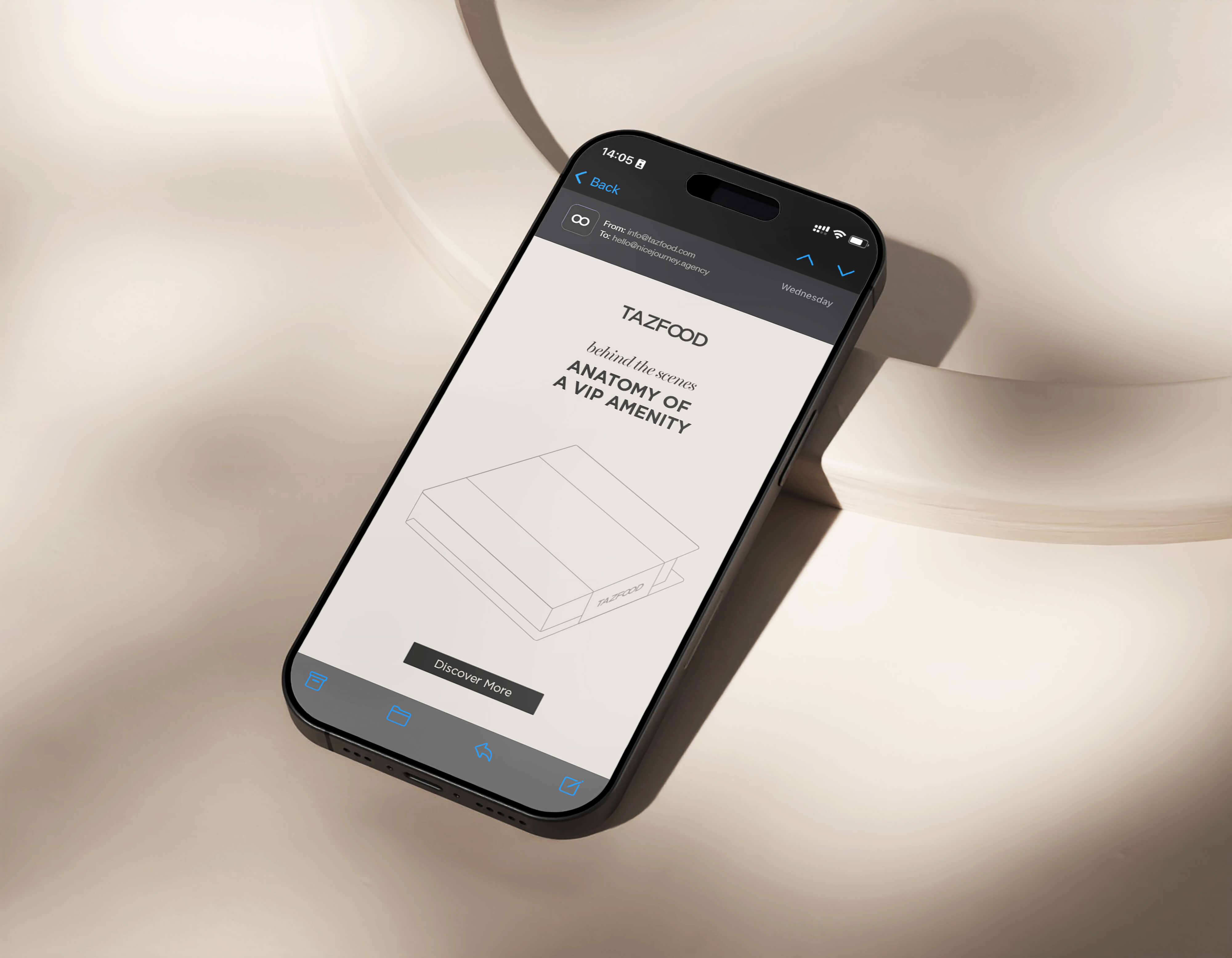 Smartphone on a beige surface displaying an email from TAZFOOD titled 'Behind the scenes anatomy of a VIP amenity' with a simple line drawing of a box and a 'Discover More' button.