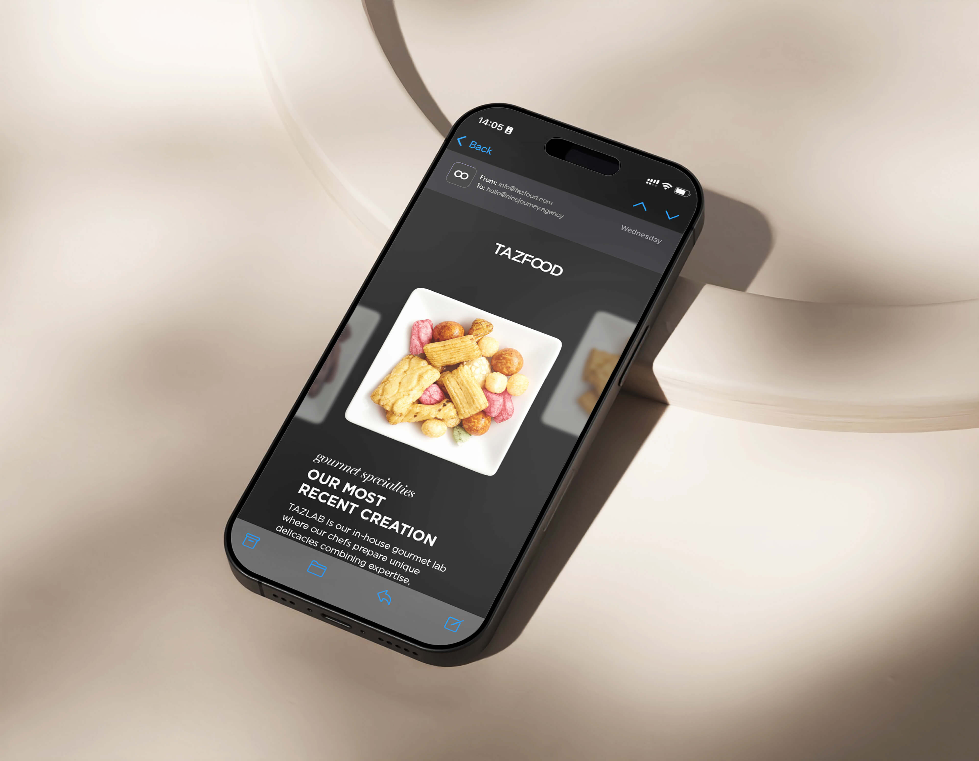 Smartphone resting on a beige surface displaying an email from TAZFOOD featuring a photo of assorted gourmet snacks on a white square plate with text about TAZLAB, the in-house gourmet lab creating unique delicacies.