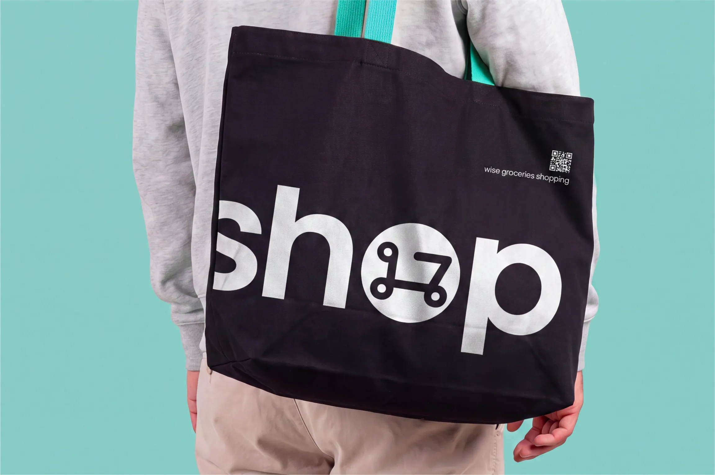 Person wearing a light gray sweatshirt and beige pants holding a black tote bag with turquoise handles, featuring the word 'shop' in large white letters with a stylized shopping cart icon inside the letter 'o' and small text that reads 'wise groceries shopping' near a QR code.