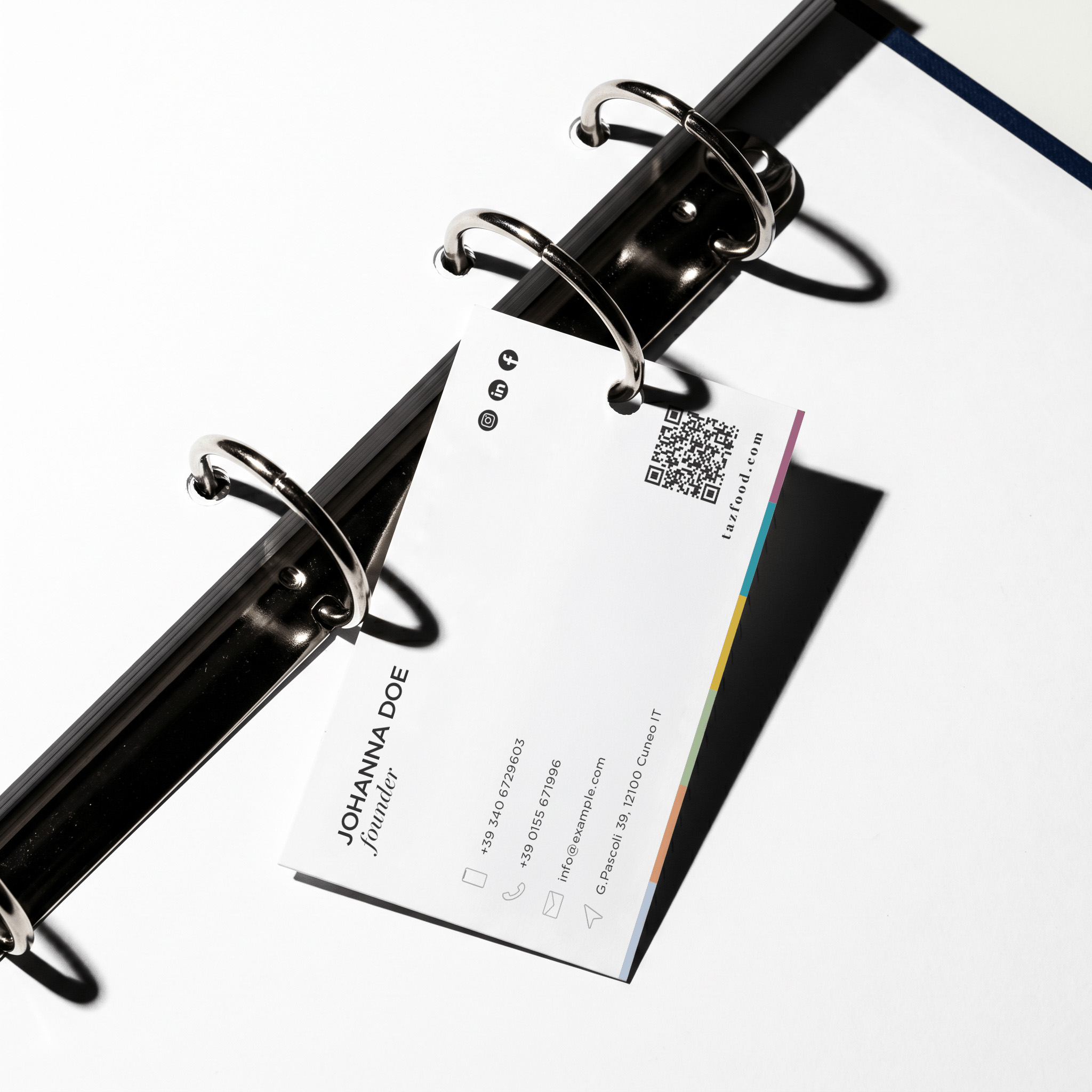 Open black metal binder ring holding a white business card with contact details for Johanna Doe, founder, including phone numbers, email, and address, with social media icons and a QR code, on a white background.