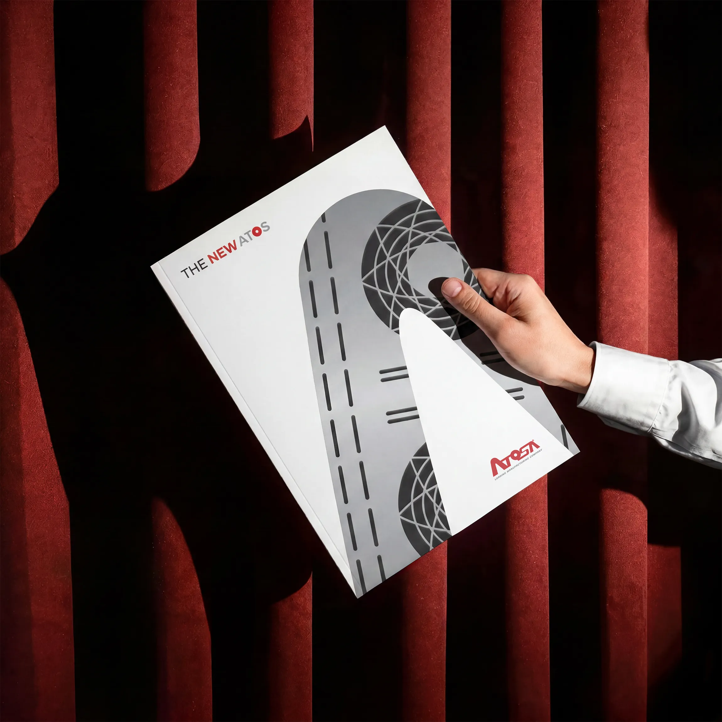 Hand in white shirt holding a magazine with text reading 'THE NEW ATOS' over a red textured curtain background.